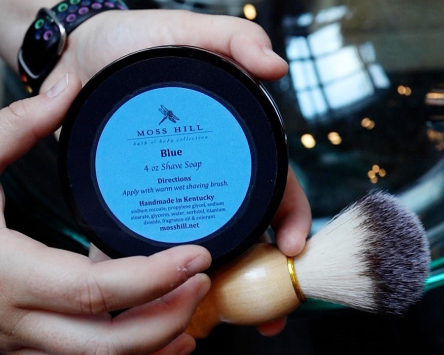 A black lid with a blue label saying 'Moss Hill Bath & Body Collection', Blue 4 oz Shave Soap, Handmade in Kentucky. Being held with a shave brush next to it.