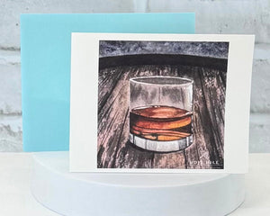 Greeting card with a glass of whiskey on a wooden surface, displayed on a white stand. That represents the Bathe in Bourbon scent collection.