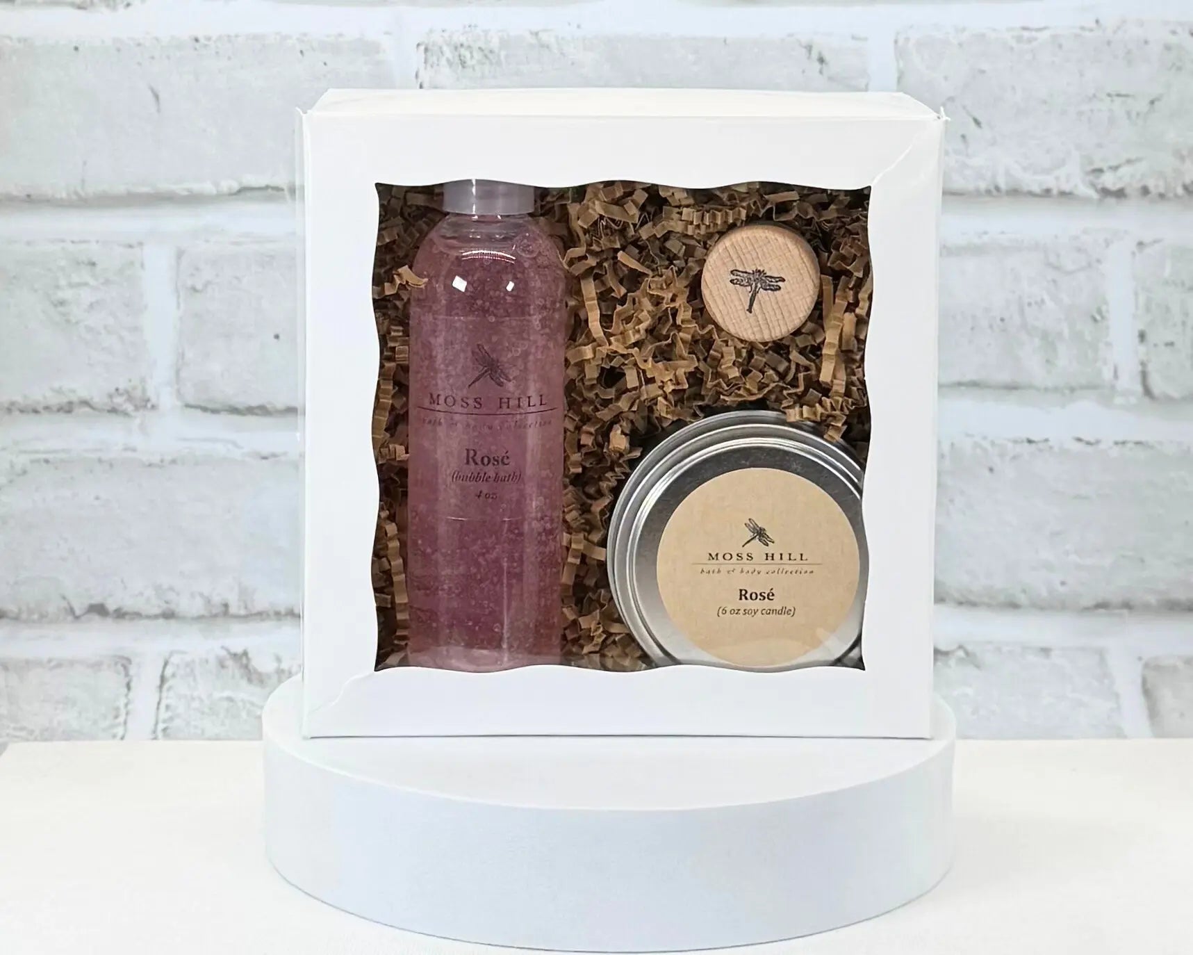Moss Hill Rose body care gift set with handmade bubble bath and a soy candle in a white box against a white brick wall.