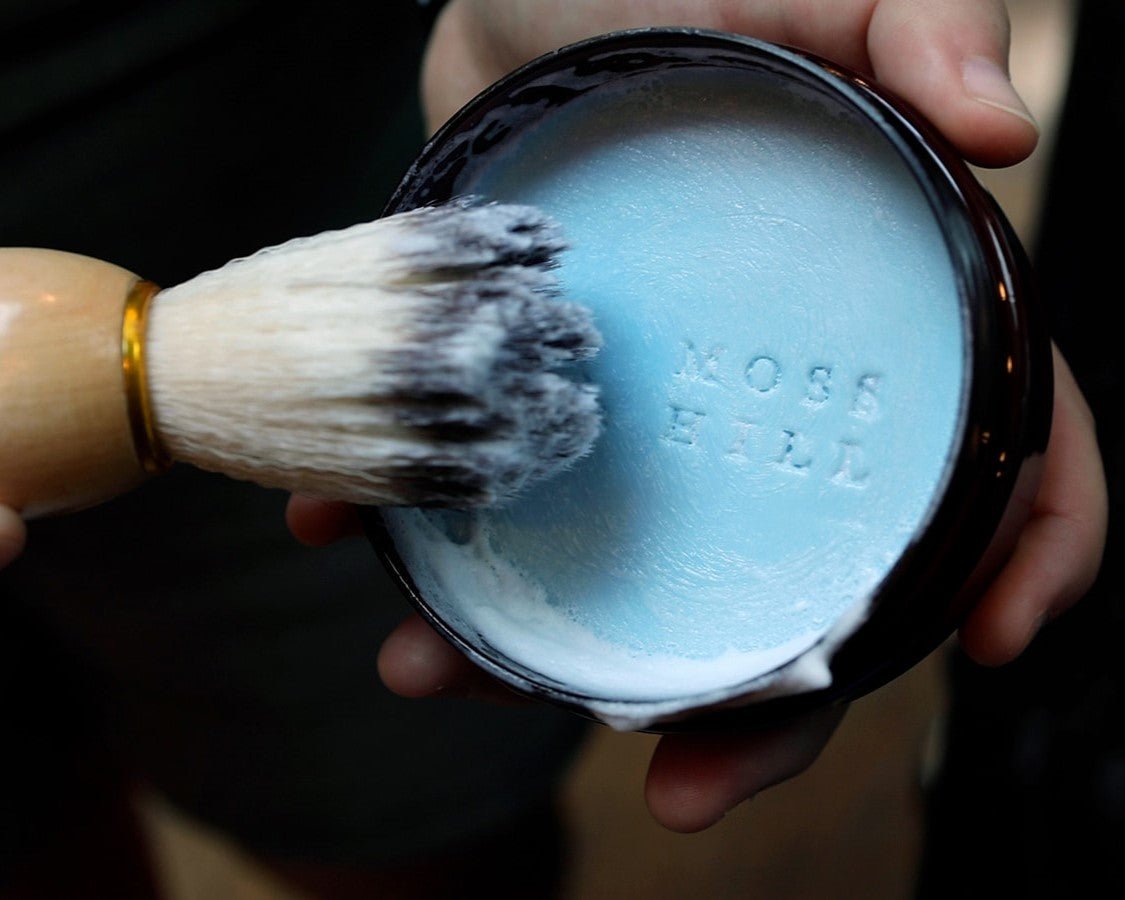 A shave brush held off to the side and shaving cream in a container lathered up with white suds.
