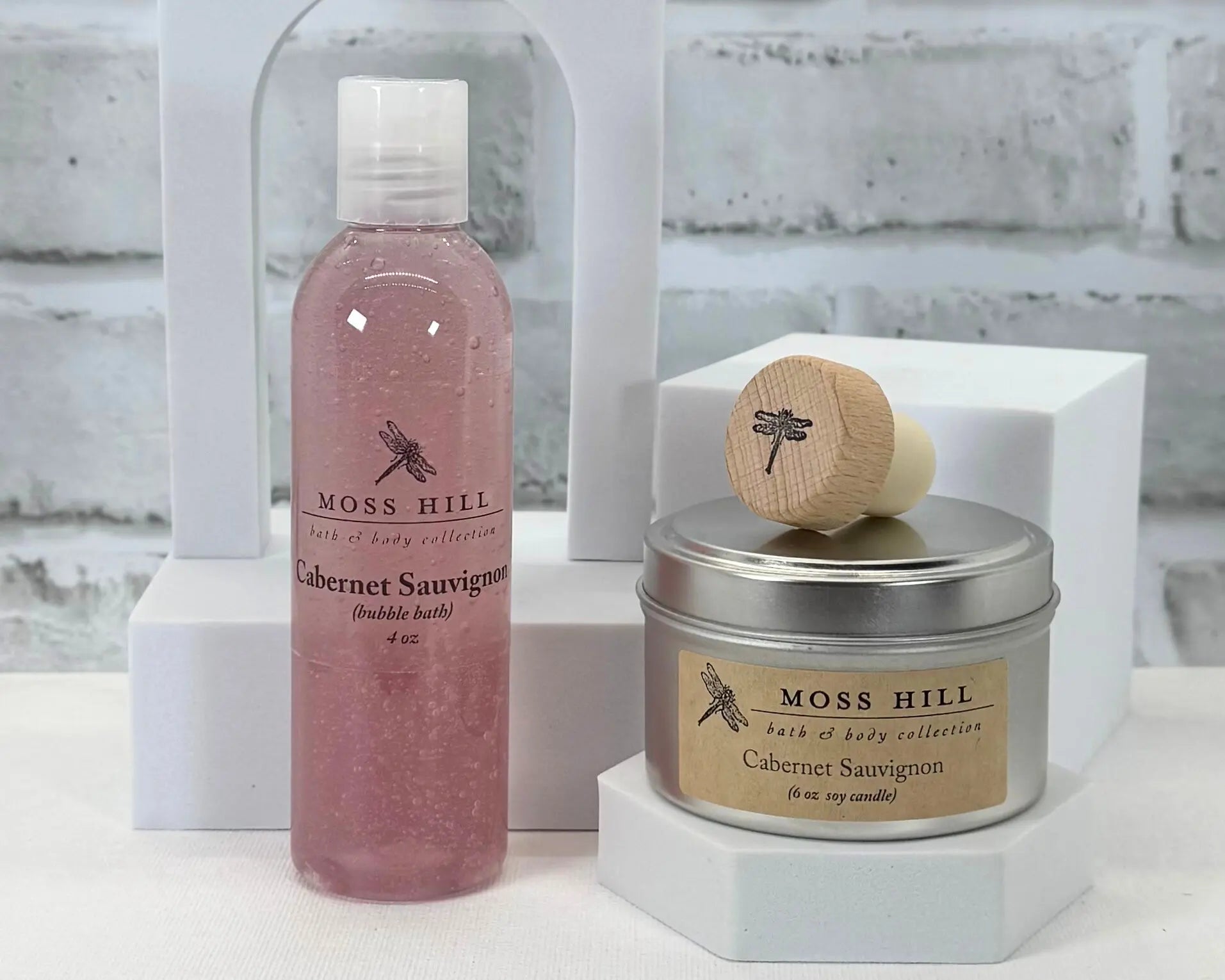 A Moss Hill body care gift set with handmade bubble bath,  soy candle, and wine cork in the scent Cabernet Sauvignon. Packaged in a white box on a white brick background.