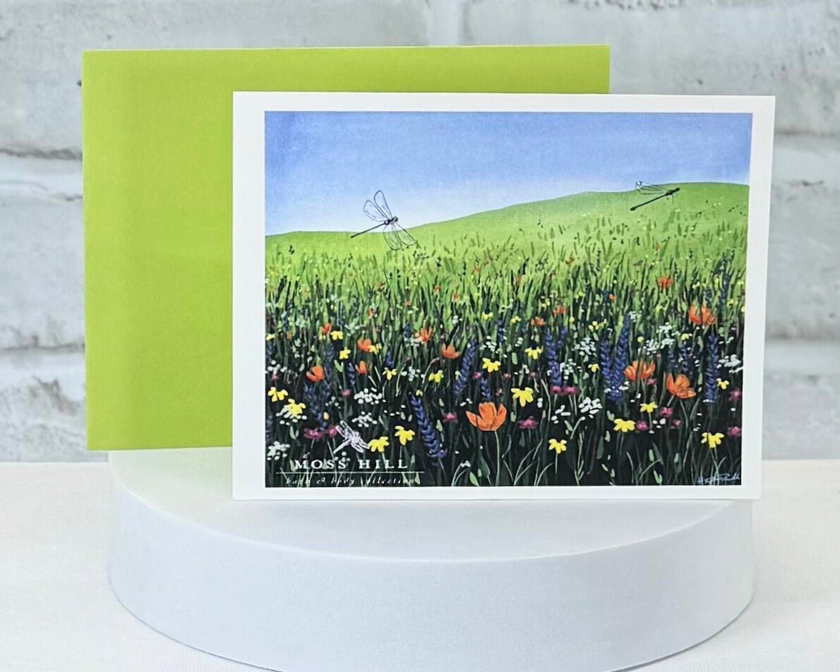 Greeting card with a field of flowers and dragonfly design on a white stand. Showcasing the scent Meadow. Which has handmade bath and body products for purchase.