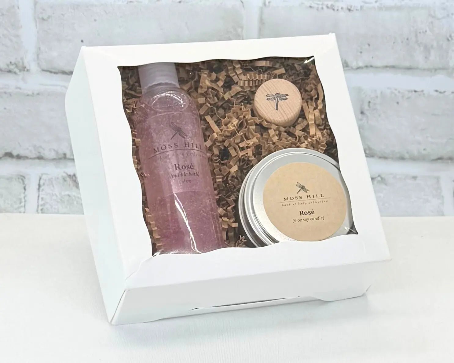 A Moss Hill body care gift set with handmade bubble bath,  soy candle, and wine cork in the scent Rose. Packaged in a white box on a white brick background.